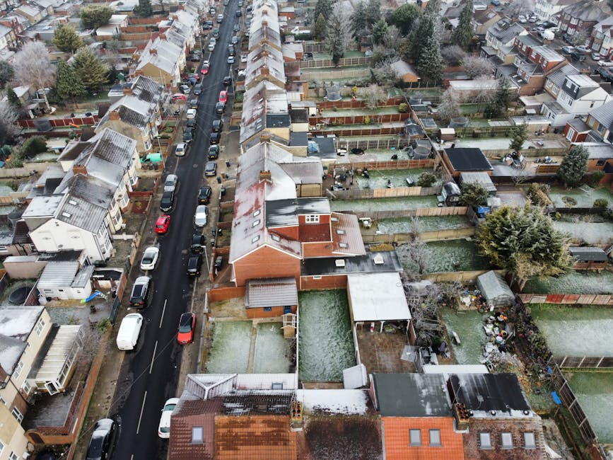 An aerial view of a residential neighborhood showing a row of terraced houses lining a narrow street, with parked cars on both sides. The image captures backyards and garden spaces behind the houses, 
